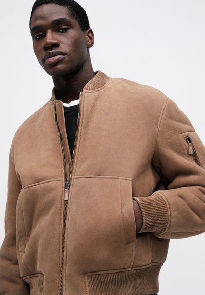 Egy férfi bézs velúr bombázó dzsekit visel, bordás mandzsettával és aljjal, cipzáros elülső része van, és az egyik kezét zsebébe dugja, háttérben egy sima fehér háttér.