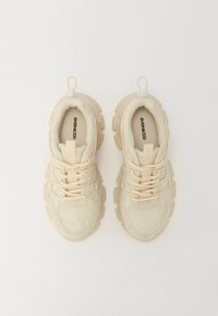 Beige athletic shoes with mesh sides, textured rubber soles, and round pull tabs. Laces are light tan; logo is printed in black inside.