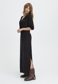 Rochie maxi neagră dintr-un material moale, cu decolteu adânc în V, mâneci bufante, fante laterale, asortată cu botine maro, dotate cu inel din metal.