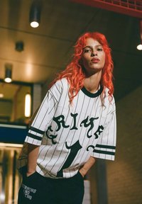 Jersey de béisbol oversize blanca con rayas verticales negras, con letras y acentos en negro en negrita, combinado con pantalones cortos negros.