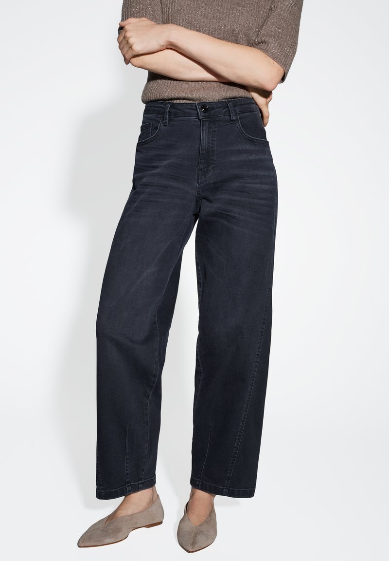 Jeans en denim à taille haute, de couleur bleu foncé, avec une coupe décontractée. Présente des poches avant et des détails de coutures subtils. Associée à des ballerines en suédine grise.