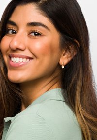 Guldörhängen i form av ringar med hjärtan, bärs av en person med långt brunt hår, som ler mjukt och är klädd i en ljusgrön skjortblus.