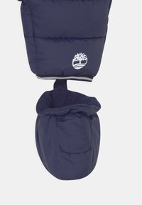 Valkoinen puuvillan vuoraama tummansininen eristetty vauvantumppu, joka on kiinnitetty samasta kankaasta valmistetun takin hihaan, jossa on fleecevuori ja valkoinen puun logo hihassa.