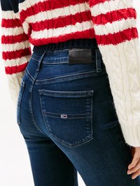 Tamni plavi traperice imaju visoki struk i oznaku s logotipom na stražnjoj strani. Model nosi pleteni pulover s bijelim i crvenim prugama.