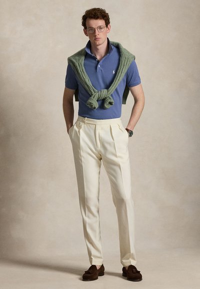 Jeune homme portant des lunettes, un polo bleu, un pantalon crème, des mocassins foncés et un pull vert posé sur les épaules, sur un arrière-plan uni.
