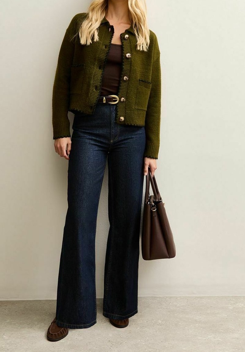 Femme portant un cardigan vert boutonné, un haut noir, un jean foncé taille haute, tenant un sac à main marron, debout contre un mur uni.