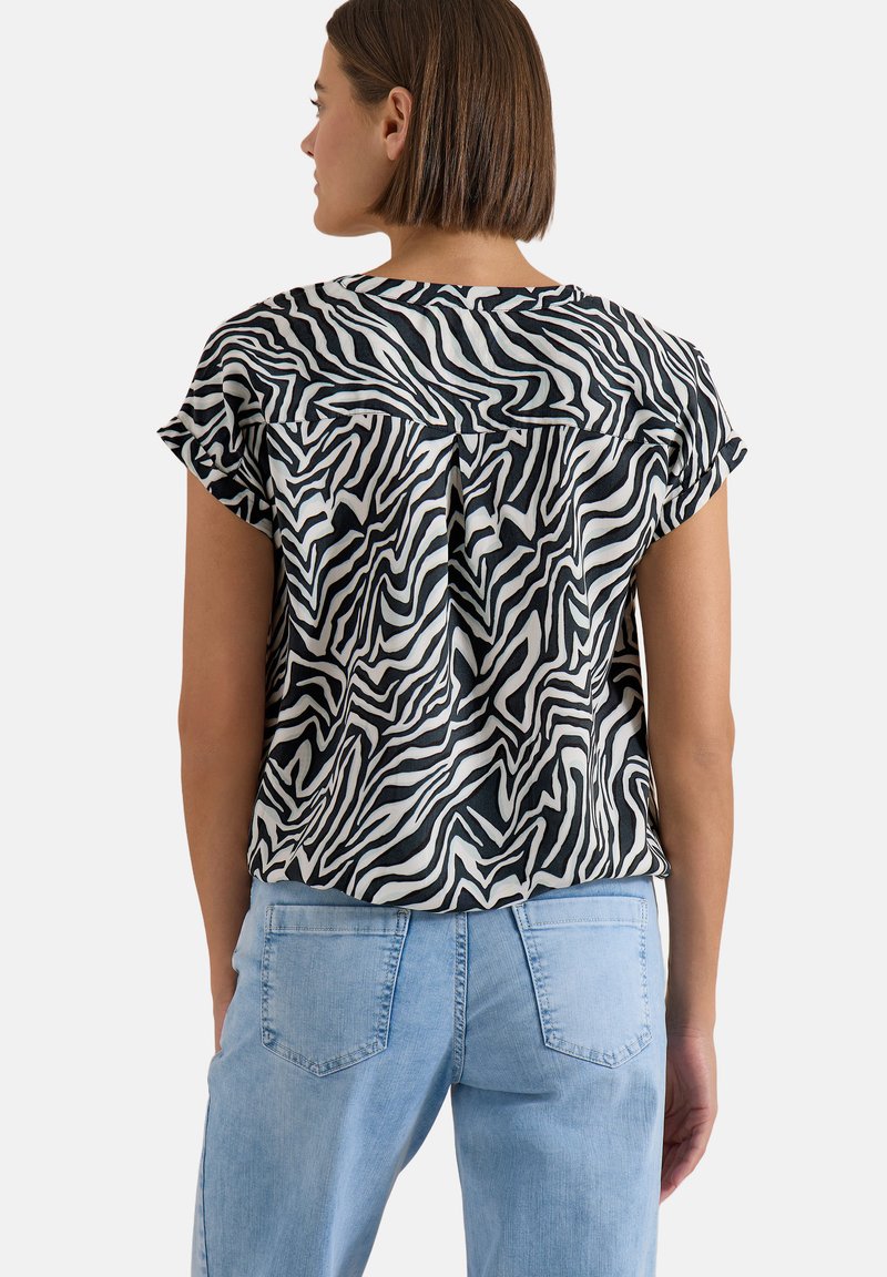 Femme aux cheveux courts et bruns portant un chemisier à manches courtes imprimé zèbre noir et blanc et un jean bleu clair, debout, de dos.