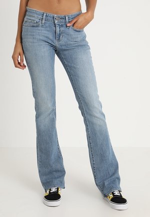 Kobieta w jasnoniebieskich dzwonowych dżinsach i czarno-żółtych tenisówkach, stojąca z jedną ręką w kieszeni na białym tle.