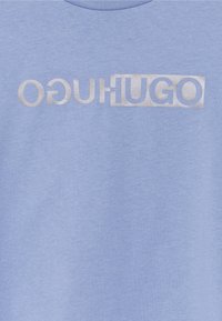 T-shirt en coton bleu avec un grand logo argenté affichant le mot "HUGO" dans un design rectangulaire. Texture douce et col rond.