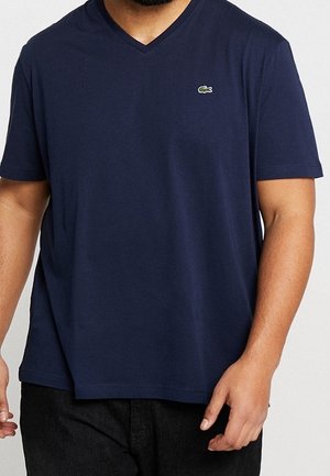 Homme portant un t-shirt à col en V bleu marine avec un petit logo de crocodile vert sur la poitrine gauche, et un pantalon noir.