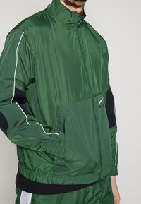 Giacca a vento verde con colletto alto, zip frontale, tasche laterali e dettagli neri. Presenta piping bianco lungo le maniche.