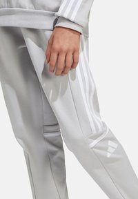 Helle graue Sweatpants mit weißen Streifen an den Seiten und Logo. Sie haben eine weiche Textur und ein tapered Design, das einen lässigen Stil unterstreicht.