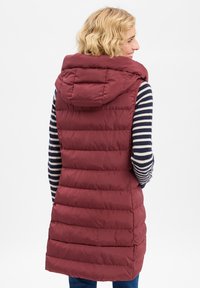 Bordeauxrote wattierte Weste mit Kapuze, mit einem gesteppten Design und einer leicht verlängerten Form. Wird über einem marineblauen und weißen gestreiften Langarmshirt getragen.