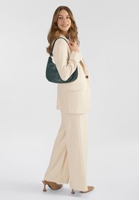 Mujer con traje crema y tacones marrón claro sosteniendo un bolso de hombro verde oscuro, sonriendo y mirando hacia un lado contra un fondo blanco liso.