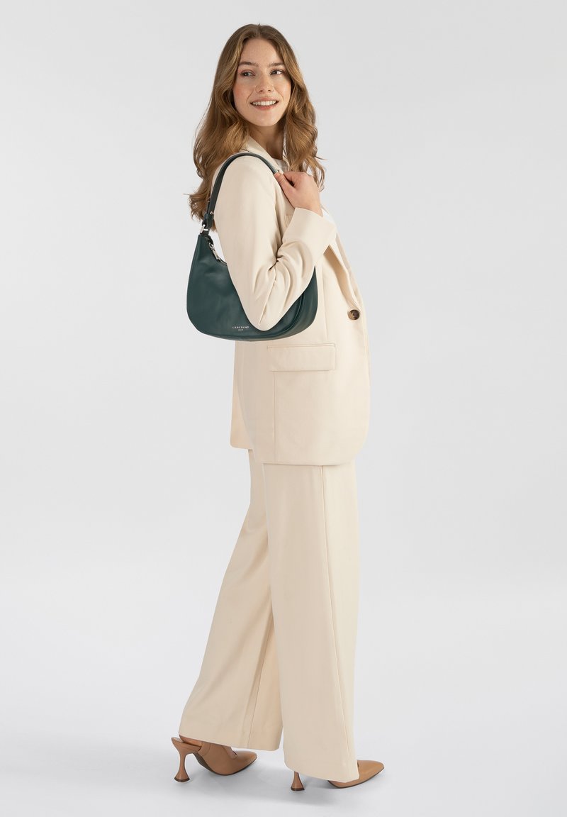 Mujer con traje crema y tacones marrón claro sosteniendo un bolso de hombro verde oscuro, sonriendo y mirando hacia un lado contra un fondo blanco liso.