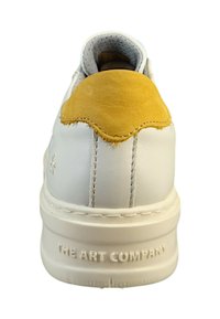 Zapatilla blanca con una lengüeta de talón de ante amarillo, cuerpo de cuero texturizado y costuras visibles. Presenta el logotipo grabado en la suela.