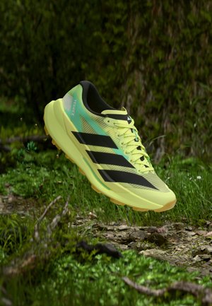 Ανοιχτό πράσινο παπούτσι adidas Terrex για τρέξιμο σε μονοπάτια με μαύρες ρίγες, αιωρούμενο πάνω από πετρώδες δάσος.