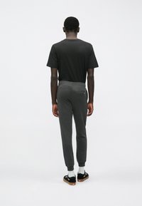 Man die met zijn rug naar de camera staat, draagt een zwart T-shirt, donkergrijze joggingsbroek met achterzak, witte sokken en zwarte sneakers met gumrubberen zolen.