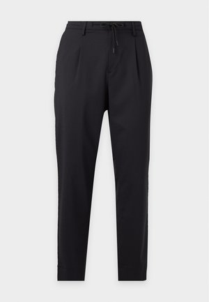Jonge man met een zwarte trui met witte "ALPES" tekst en coördinaten, zwarte broek, witte sokken en zwarte sneakers, staand tegen een witte achtergrond.