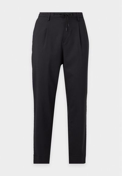 Pantalon noir fuselé avec plis, passants de ceinture et cordon de serrage à l'avant sur fond clair uni.