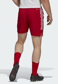 Man in rode sportshorts, rode sokken en zwarte voetbalschoenen, die weglopen met één voet omhoog, tegen een effen lichte achtergrond.