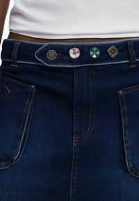 Jupe en denim de couleur bleu foncé, dotée de quatre boutons uniques à la taille et d'une couture jaune contrastante sur les poches avant.