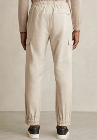 Pantalons cargo beiges avec une taille élastique, dotés de deux poches latérales, de poignets élastiques et d'une texture de tissu lisse. Une paire de baskets noires en dessous.