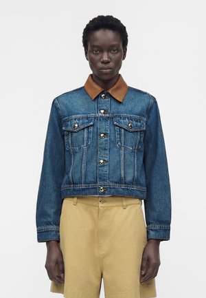 Osoba w niebieskiej kurtce dżinsowej z brązowym skórzanym kołnierzem i beżowych szortach, stojąca na jednolitym jasnym tle.