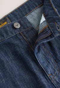Blugi din denim cu o nuanță albastră închisă, având fermoar la deschidere, accesorii din metal argintiu și cusături contrastante de culoare portocalie. Foderul interior este vizibil.