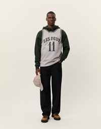 Halli korvpallil on hall võrkmaterjal, millel on tumeroheline tekst "LES DEUX 11". See on ühendatud rohelise kapuutsiga sviitriga, tumedate pükste ja triibulise mütsiga.