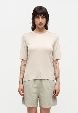 Femme aux cheveux foncés portant un haut beige à manches courtes et un short gris clair, debout devant un fond blanc uni.