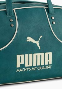 Tyrkisblå Puma-håndtaske med hvidt logo og teksten "MACHT'S MIT QUALITÄT" på tysk, med hvide kanter og sølvnitter nær håndtagene.