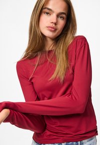 Jonge vrouw met lang lichtbruin haar, gekleed in een aansluitend rood langärmshirt en lichtblauwe jeans, tegen een witte achtergrond.