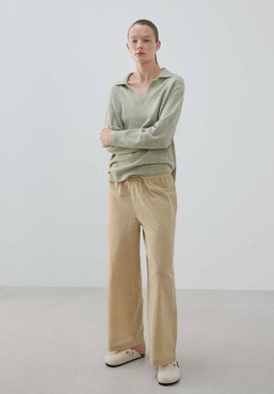 Joven mujer de pie con los brazos cruzados, vistiendo una camiseta de manga larga verde claro, pantalones anchos beige y zapatos tipo mocasín de color crema.