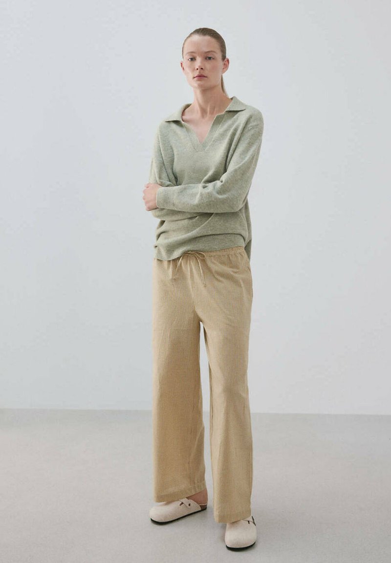 Jeune femme debout, les bras croisés, portant un haut à manches longues vert clair, un pantalon large beige et des chaussures slip-on couleur crème.
