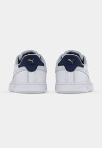Pár bílých tenisek Puma s tmavě modrými patními záložkami a zlatými logy Puma, pohled zezadu na jednobarevném bílém pozadí.