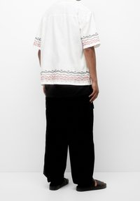 Hvid kortærmet skjorte med rød og sort geometrisk broderi nederst, parret med løse sorte bukser og mørke sandaler.