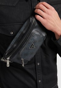 Sac banane noir et bleu avec un motif, une surface texturée, une fermeture éclair argentée et un logo triangulaire. Porté en travers de la poitrine.