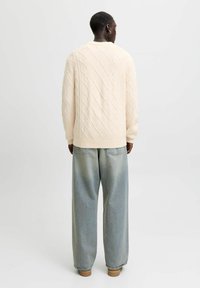 Cremefarbener Pullover mit einem Rautenmuster aus Zopfmuster, rundem Halsausschnitt und gerippten Bündchen, kombiniert mit hellblauen, weit geschnittenen Jeans.
