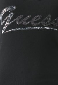 Top morbido a righe nere con logo "Guess" in strass, con effetto scintillante e design aderente.