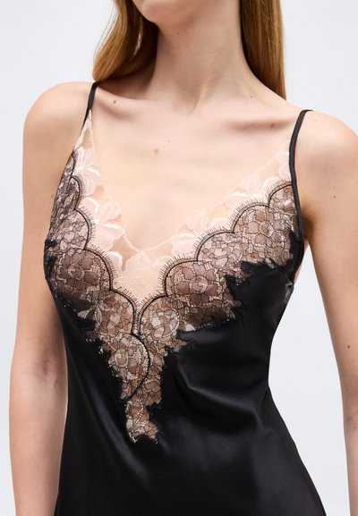 Γυναίκα που φοράει μαύρο σατέν φόρεμα με λεπτές τιράντες και διαφανή μπεζ και μαύρη δαντέλα με floral σχέδιο κατά μήκος του βαθιού V-λαιμού.