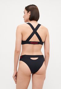 Črn bikinij komplet s križnim trakom. Vključuje logotipne poudarke v rdeči barvi na zadnji strani zgornjega dela in pasu. Zgibana tkanina.
