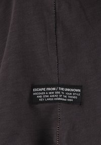 Tejido negro con un acabado texturizado que presenta una etiqueta. El texto de la etiqueta incluye "ESCAPAR DE / LO DESCONOCIDO," con un detalle de costura blanca.