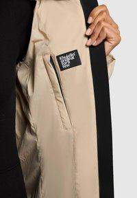 Hand holding open beige jacket, showing inner pocket with black rectangular label displaying white text "Khujo original for a few."