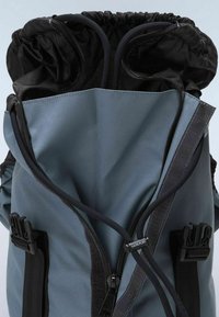 Interior de mochila de tela gris con parte superior de cordón negra, cierre resistente y correas ajustables negras. Presenta un acabado texturizado y etiqueta de marca.