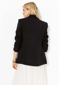 Blazer noir à manches longues, de forme cintrée, avec une texture subtile. Porté sur une jupe en dentelle blanche, mettant en avant un design épuré et minimaliste.