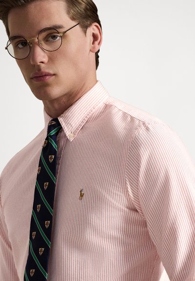 Ριγέ ροζ πουκάμισο με κουμπιά και κλασικό γιακά, με μικρό κεντητό λογότυπο στο στήθος, σε συνδυασμό με μπλε γραβάτα διακοσμημένη με ασπίδες.