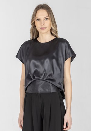 Kobieta o blond włosach do ramion, ubrana w błyszczącą czarną bluzkę z krótkim rękawem i szerokie czarne spodnie, stojąca na tle jednolitego jasnego tła.