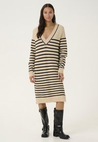 Robe pull en beige avec des rayures horizontales noires, décolleté en V et manches longues. Assortie avec des bottes noires jusqu'aux genoux.