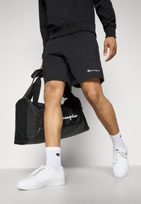 Black sports shorts with logo, paired with white sneakers and socks. A black duffel bag with white logo accents is held at the side.